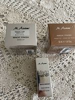 Three boxed M. Asam makeup products on a lace background: two Magic Finish foundation boxes and one Magic Mango lip gloss box.
