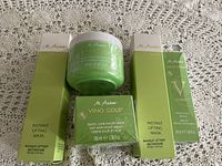 Five M.Asam Vino Gold skin care products displayed on lace background include a green jar and four green and white boxes.