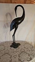 Front-side angle view of the black metal heron statue standing on a patterned fabric surface with white backdrop.