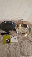 View of green ridged frying pan and round metal steamer insert on a table along with recipe booklet and instruction manual.