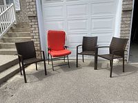 Wide view showing three brown plastic wicker patio chairs with arms and one black folding chair with a red cushion arranged outdoors in front of a garage door.