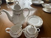 Photo showing teapot with lid, creamer, sugar bowl with lid, and seven stacked dessert plates on wooden table, all part of Royal Doulton tea service.