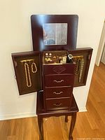 Opening jewellery box showing mirror, side doors with hanging necklaces, and compartments with assorted costume jewellery including earrings, brooches, and a wristwatch.