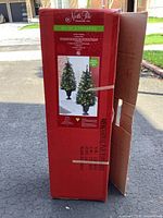 Red boxed packaging featuring image of two illuminated artificial Christmas tree topiaries standing on black urn bases.