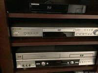 Photo showing three electronic media players stacked on shelves: Panasonic DVD player, Samsung Blu-ray player, JVC VCR/DVD combo unit.