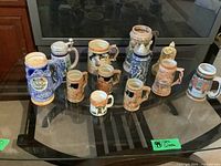 Overview of 11 German beer mugs and steins with varying heights and designs on glass table.