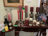 Wide view of assorted candlesticks and unused candles displayed on a wooden table including clear glass holders, ornate gold-tone holders, and painted floral pillar candles.