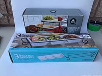 Photo of two boxed 3-section white stoneware serving trays with black metal wire stands, showing original packaging and branding.