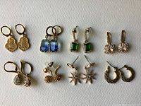 Top-down view of 8 pairs of vintage costume jewelry earrings laid out on white textured background.