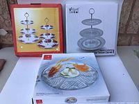 Three tiered sandwich stands and serving platter in original retail boxes displayed on white surface.