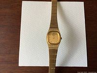 Top-down view of the gold tone ladies Seiko watch laid flat on white cardstock on wood surface showing the entire bracelet and face of the watch.