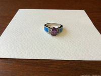 Front view of vintage silver ring showcasing purple amethyst center and blue synthetic opal side inlays on a white textured paper background.