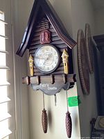 Full view of the vintage wooden chalet cuckoo clock mounted on wall next to window with pine cone weights hanging