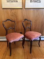 Two antique wooden balloon back chairs with dark wood frames and dusty rose upholstered seats placed side by side on a wooden floor against a wood paneled wall.