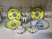 View of all items in lot arranged on kitchen counter, showing three yellow round plates, one oval pig motif plate, lidded pitcher and small teapot and sugar bowl.