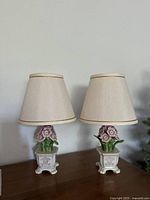 Front view of both petite antique ceramic floral table lamps with cream fabric shades on wooden surface.
