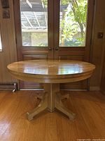 Full view of antique round wooden pedestal dining table placed on wooden floor with double glass doors in background.