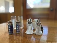 Close-up photo of two pairs of salt and pepper shakers on a wooden table including blue metal lattice and ribbed clear glass with silver tops.