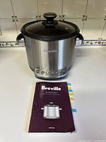 Front view of Breville Risotto Plus cooker with glass lid and original manual on white surface.