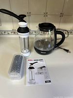 Photo showing the OXO cookie press with clear plastic cylinder and black and white handle, the box of metal disc attachments in a separate plastic container, the electric glass kettle with black handle and base, and the OXO instruction manual.