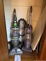 Hoover WindTunnel MaxExtract vacuum and Shark Navigator vacuum standing side by side with ironing board behind them.