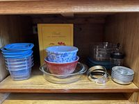 Kitchen cabinet shelf showing assorted plastic containers with blue lids, ceramic bowls (red and blue floral), clear glass baking dishes, metal ring biscuit cutters, a cockatoo ceramic container, and a yellow cookbook titled 'Almost All About Chicken'.