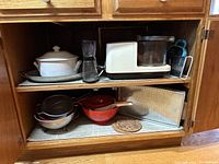 View of open cabinet showing white slow cooker, blender, white toaster, food processor, metal colanders, red cast iron cookware, glass tray, and cork trivets.