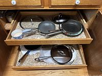Photo showing two kitchen drawers open containing multiple All-Clad pans and lids including frying pans and saucepans.