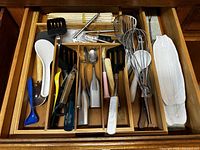 Top-down photo of an open kitchen drawer filled with various large kitchen utensils and a white ceramic corn-shaped dish.
