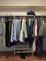 Photo of assorted men's vintage clothing on hangers including pants, shirts, jackets, and sweaters along a closet rod with shoes below.