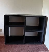 Black modular bookcase cabinet standing against a wall, showing its front with four open shelving spaces divided vertically and horizontally.