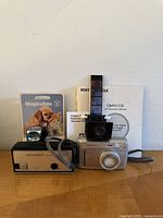 Front view of the Pentax Optio 33L digital camera, Mamiya 100 camera, flash unit, Magicubes packaging, and Pentax manuals displayed on a white surface.