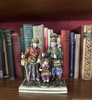 Front view of the hand-painted porcelain family figurine of beggars on a bookshelf with books in background.