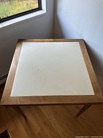 Full view of the maple folding card table showing the beige leather-like top seated in a maple wood frame on a wood floor near window.