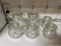 Photo showing six vintage clear glass storage jars with bubble design, three have lids, three do not, placed on a kitchen counter near a tiled backsplash with an electrical outlet.