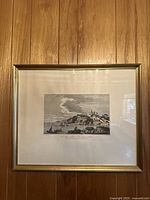 Framed vintage coastal art print with boats and village scene on wood floor background.