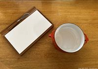 Overhead view of the teak wood cheese board with ceramic insert and cheese spreader knife next to the Dansk red enamel casserole pot interior.