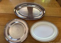 Photo of three items: oval Noritake Nerrisa china serving platter with creamy white and gold border, round silver serving tray, and oval silver serving tray with etched design