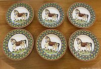 Top view of all six plates arranged in two rows on a wooden surface, showcasing the horse designs and colorful patterned borders.
