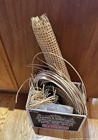 Box of vintage weaving materials including rolled cane mesh, wicker strips, and bundles of straw placed vertically in a wooden background setting.