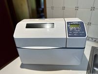 Front view of the Zojirushi breadmaker showing its white body and vented lid with control panel on the right side.