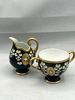 Photo of porcelain creamer and sugar bowl set with hand-painted yellow and lavender floral motifs on deep navy background, pink enamel medallions, and gilt accents.