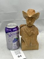 Photo showing wooden cowboy bust beside a soda can for size reference, highlighting its approx. 7.5 inch height, natural unfinished wood surface, and detailed carving work on the face and hat.