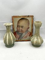 Two small crackle glaze pottery vases with a framed print of a child behind them against a white background