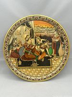 Front view of round marble plate featuring detailed Mughal harem court painting with floral border and 24k gold trim.