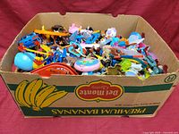 A large cardboard box filled with an assortment of colorful plastic toys including vehicles, figures, and musical toys.