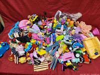 Overview of a large box filled with assorted loose toys including dolls, toy vehicles, and small playsets on a red background.