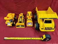 Photo shows four yellow CAT construction vehicle toys on red fabric background with measuring tape for scale.