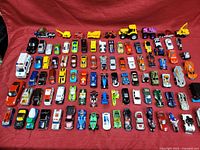 Full overview of approximately 54 diecast toy cars and vehicles spread on red fabric, displaying variety in size, color, and style.