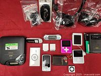 All electronic devices laid out on red surface, including various MP3 and MP4 players, iPods, Philips and Sony players, and multiple earphone sets.